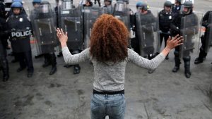 a black woman stands in the foreground, back to the camera and hands up. She faces a line of police officers in riot gear