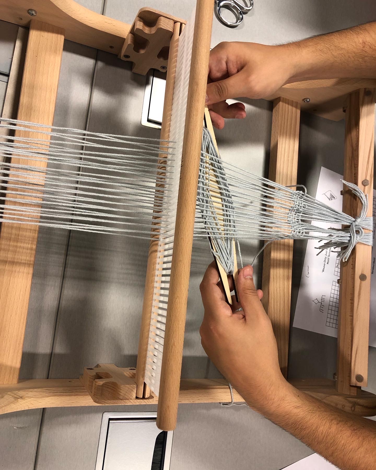 Hands guide the shuttle through the warp of the heddle loom