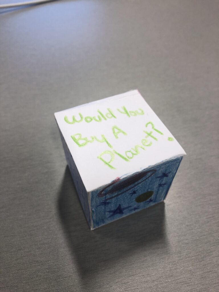 A cube made of white paper, which themaker has decorated in different colors. The top reads "Would you buy a planet?"