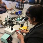 A white woman, seated, looking through magnifying lenses at the circuit she is soldering