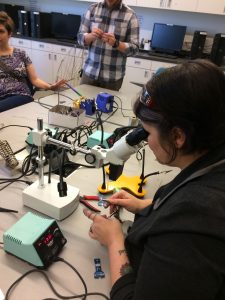 A white woman, seated, looking through magnifying lenses at the circuit she is soldering