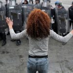a black woman stands in the foreground, back to the camera and hands up. She faces a line of police officers in riot gear