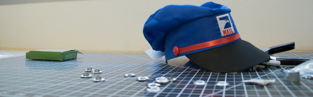 Pretend mail carrier hat sitting on a cutting mat, surrounded by grommets